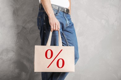 Man holding paper shopping bag with percent sign near grey wall, closeup. Discount concept