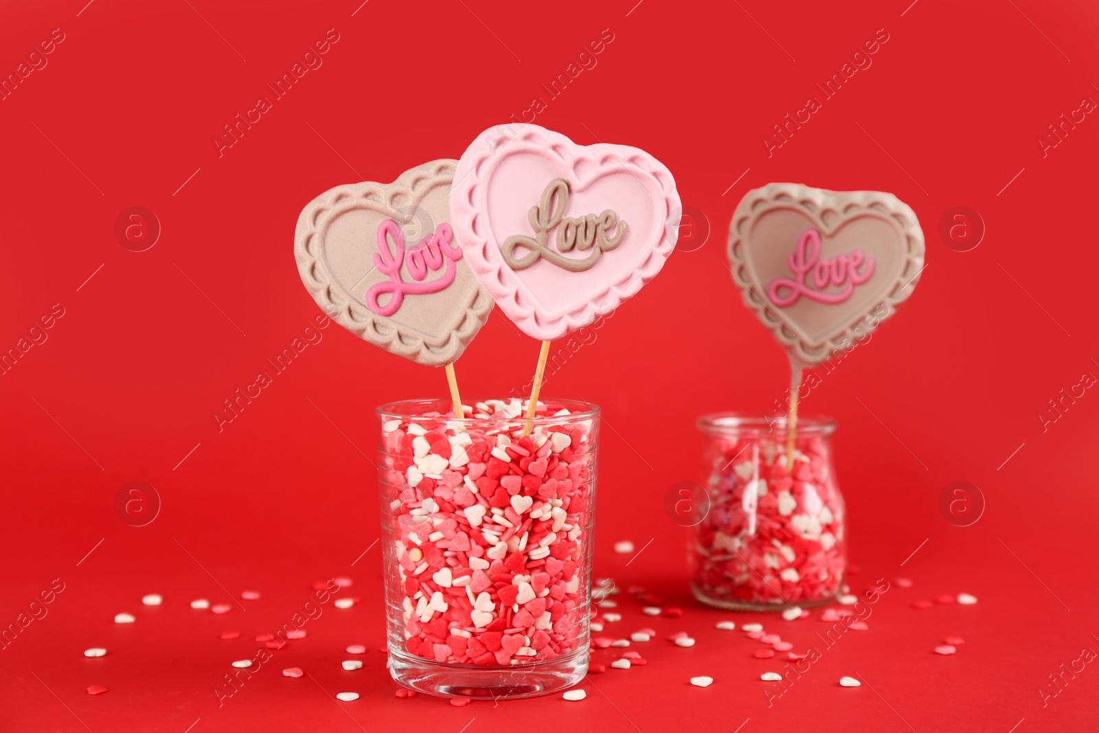 Photo of Heart shaped lollipops made of chocolate with sprinkles in glasses on red background