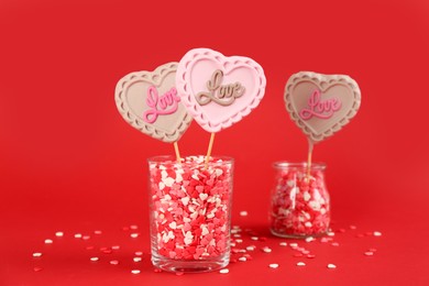 Heart shaped lollipops made of chocolate with sprinkles in glasses on red background