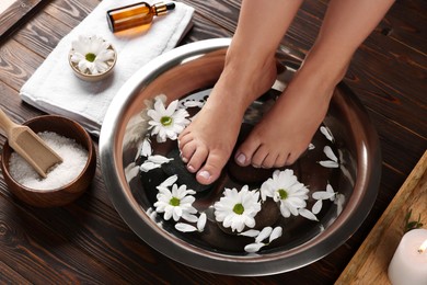 Woman soaking her feet in bowl with water, stones and flowers on wooden floor, closeup. Pedicure procedure