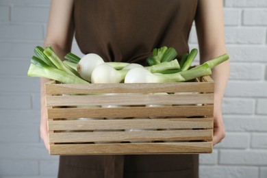 Woman holding wooden crate with green spring onions near white brick wall, closeup