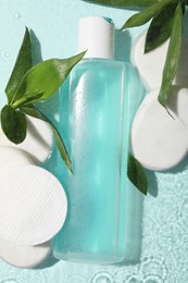 Photo of Wet bottle of micellar water, cotton pads and green twigs on light blue background, flat lay
