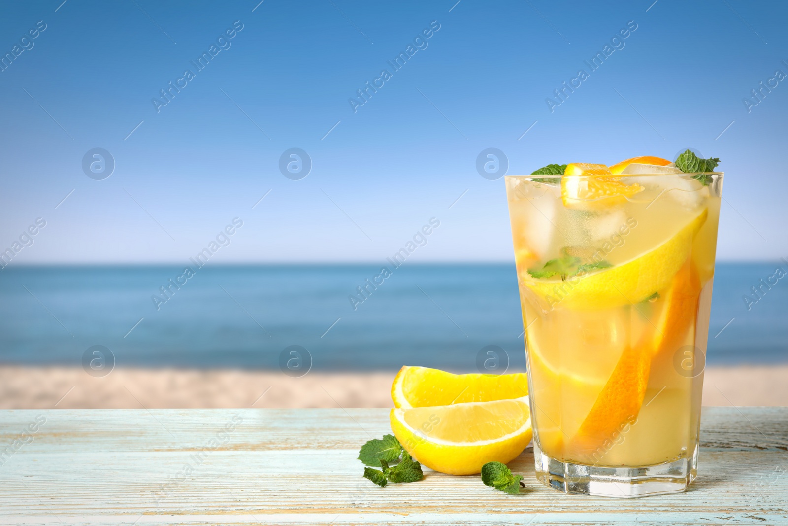 Image of Citrus lemonade with ice cubes on wooden table near sea, space for text