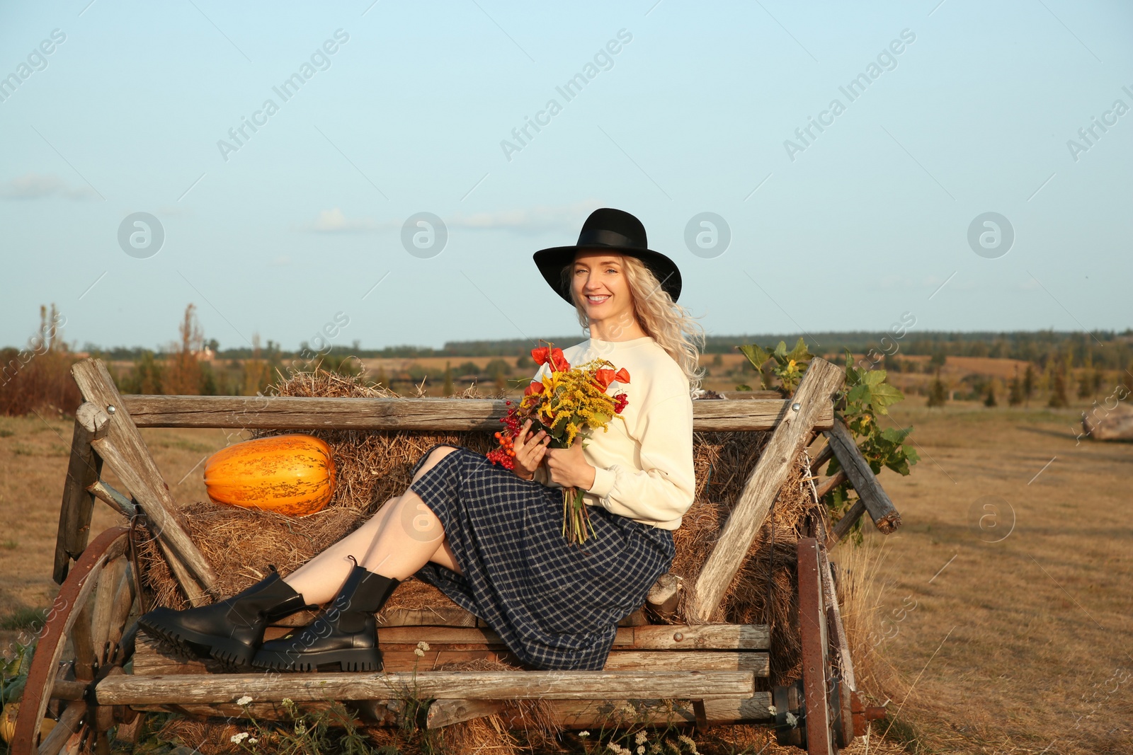 Photo of Beautiful woman with bouquet sitting on wooden cart with pumpkin and hay in field. Autumn season