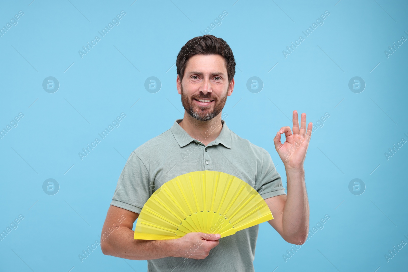 Photo of Happy man holding hand fan and showing ok gesture on light blue background