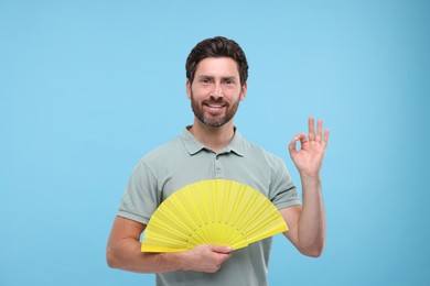 Happy man holding hand fan and showing ok gesture on light blue background