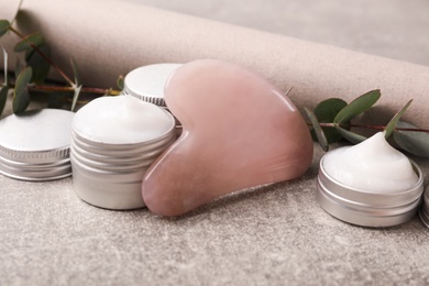 Photo of Rose quartz gua sha tool, jars of cream and eucalyptus branches on grey table, closeup