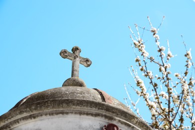 Photo of Roof of old chapel with stone cross near blossoming tree against blue sky, closeup
