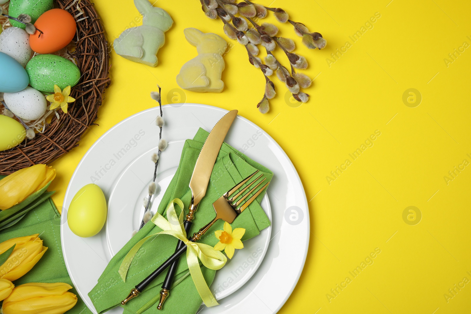 Photo of Festive table setting with painted eggs and tulips on yellow background, flat lay. Easter celebration