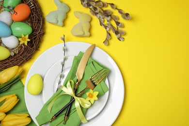 Festive table setting with painted eggs and tulips on yellow background, flat lay. Easter celebration