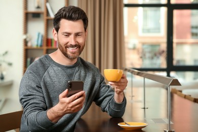 Photo of Handsome man with cup of coffee using smartphone at table in cafe