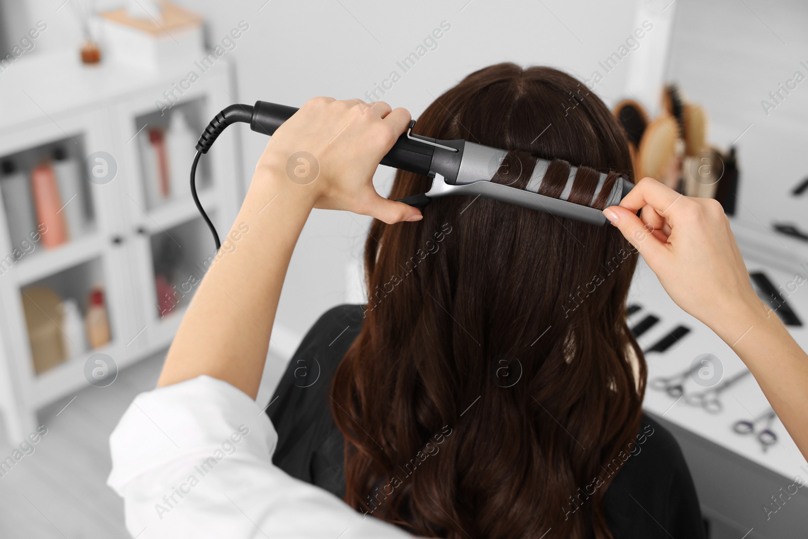 Photo of Hairdresser working with client using curling hair iron in salon, closeup