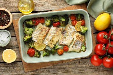 Photo of Pieces of delicious baked cod with vegetables, lemon and spices in dish on wooden table