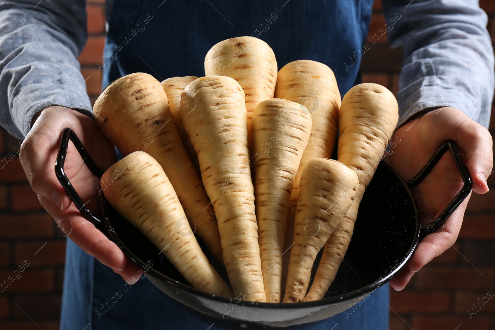 Photo of Woman holding bowl with fresh ripe parsnips near red brick wall, closeup