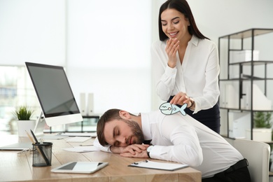 Young woman sticking paper fish to colleague's back while he sleeping in office. Funny joke