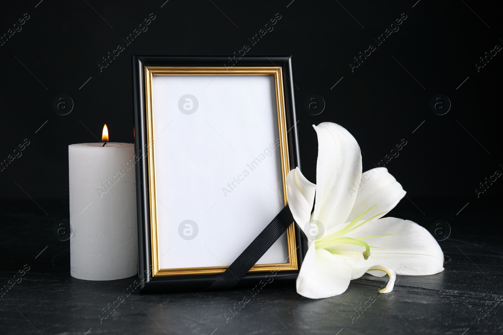 Photo of Funeral photo frame with ribbon, white lily and candle on black table against dark background. Space for design