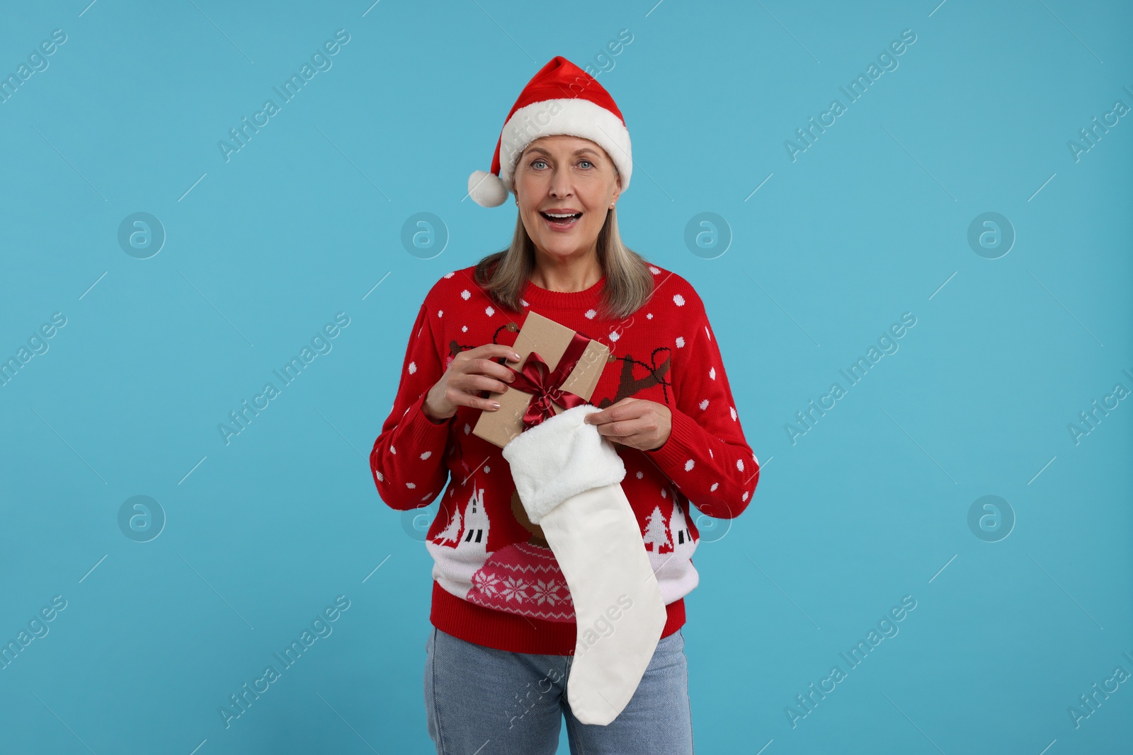 Photo of Surprised senior woman in Christmas sweater and Santa hat taking gift from stocking on light blue background