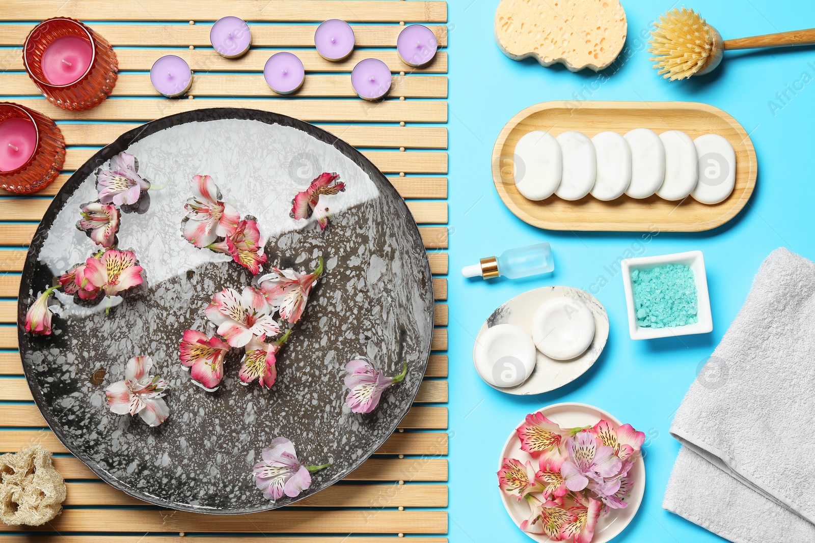 Photo of Bowl of water with flowers and different spa supplies on light blue background, flat lay