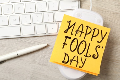 Computer mouse with Happy Fools' Day note near keyboard on wooden table, above view