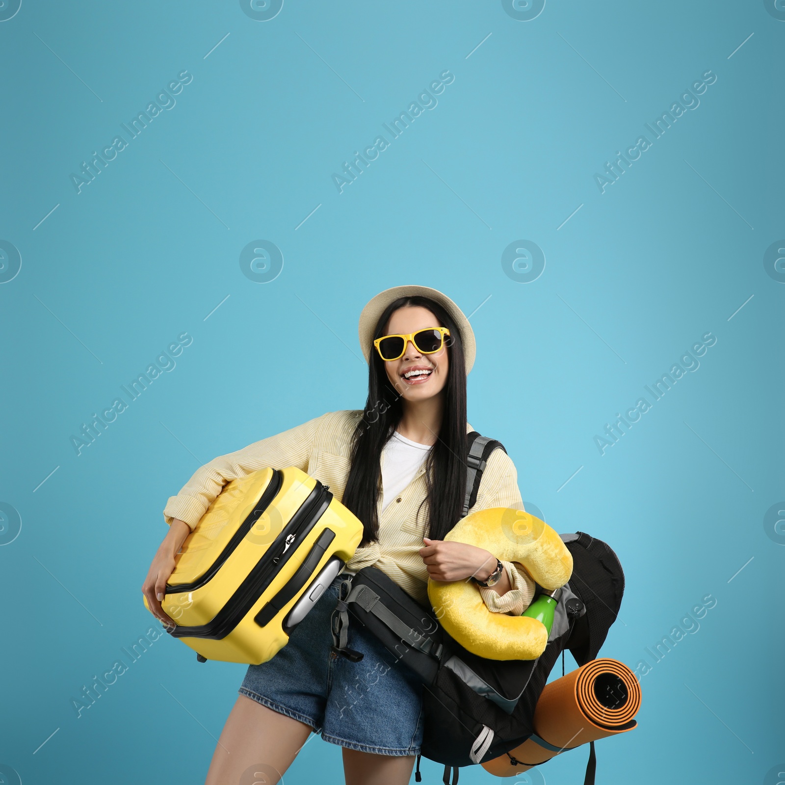 Photo of Happy female tourist with backpack, suitcase and travel pillow on light blue background