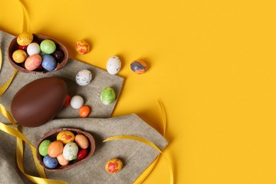 Whole and halves of chocolate eggs with colorful candies on yellow background, flat lay. Space for text