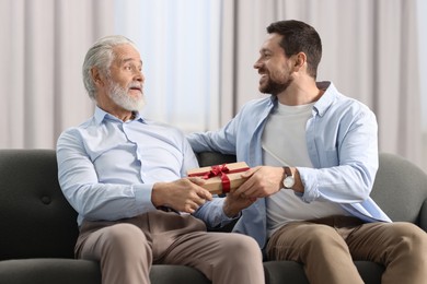 Photo of Son giving gift box to his dad on sofa at home