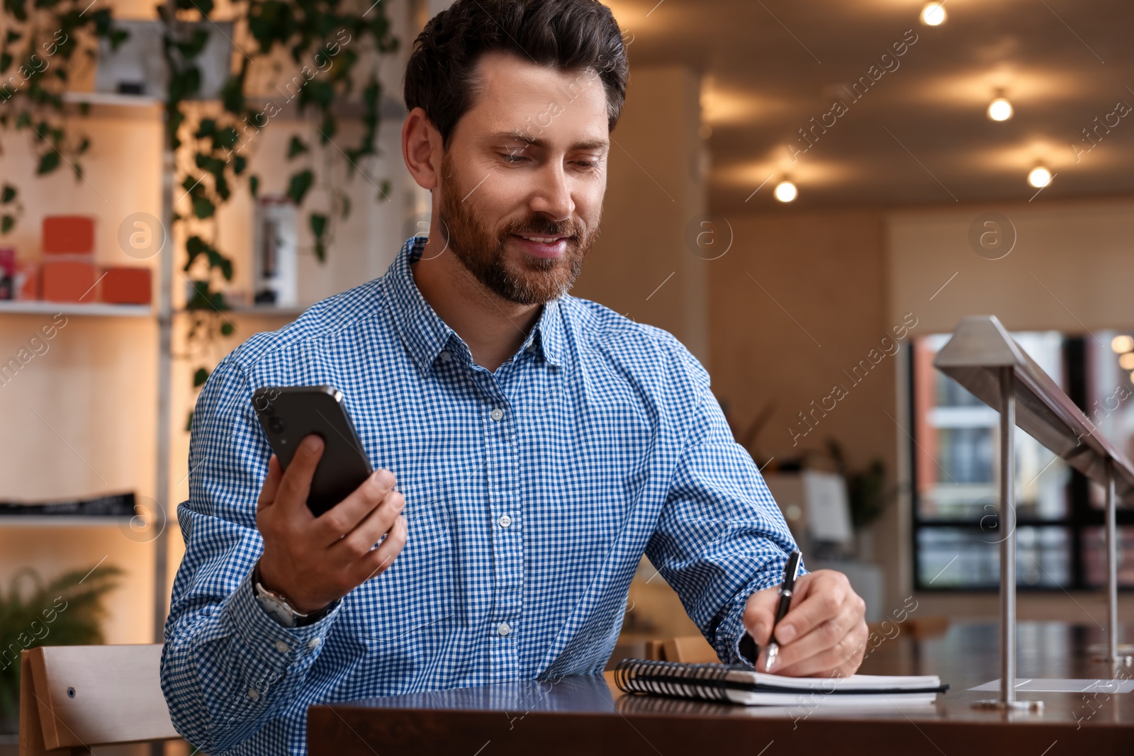 Photo of Handsome man with smartphone writing something in notebook at table in cafe