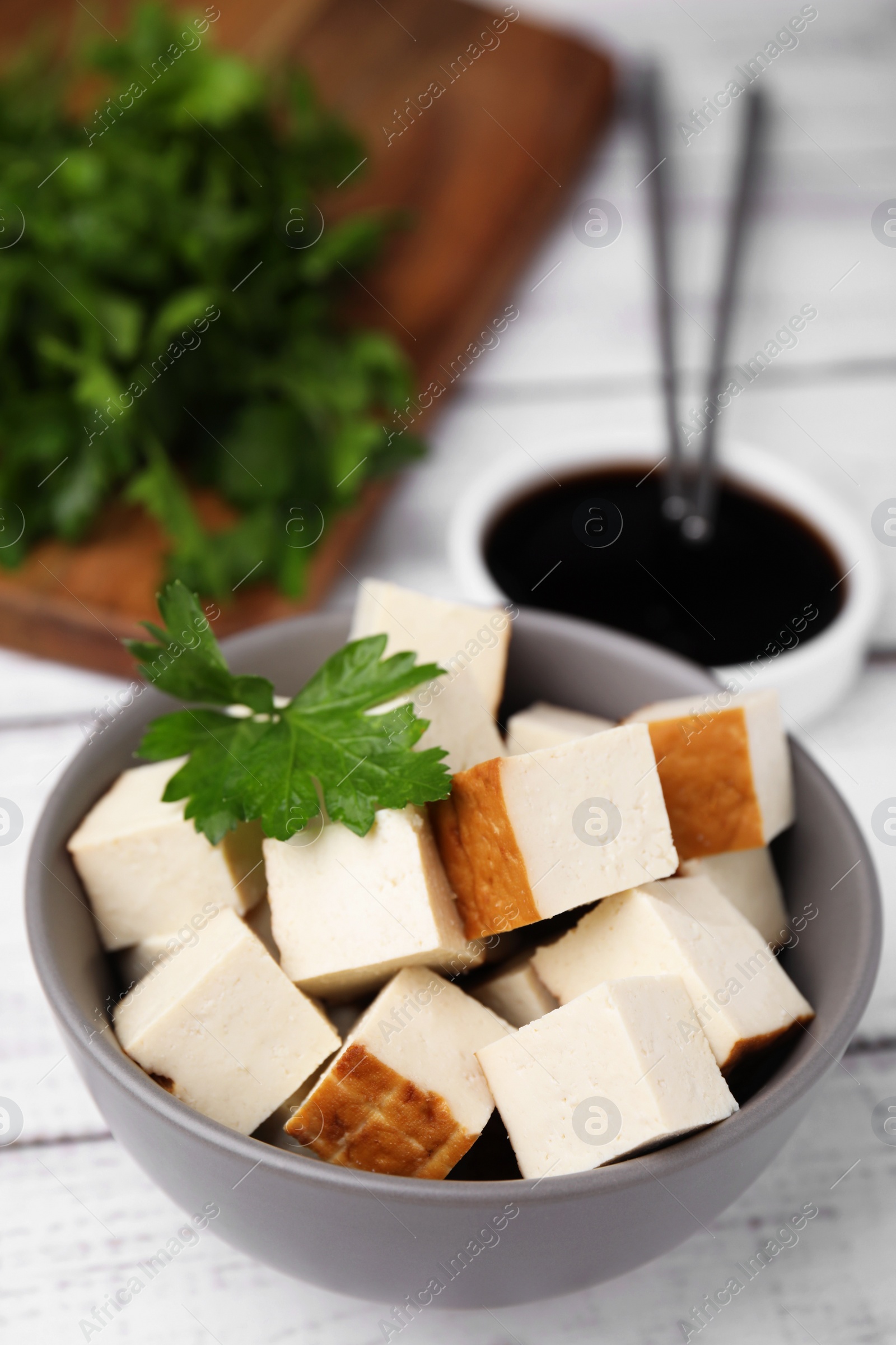 Photo of Bowl of smoked tofu cubes, soy sauce and parsley on white wooden table, closeup