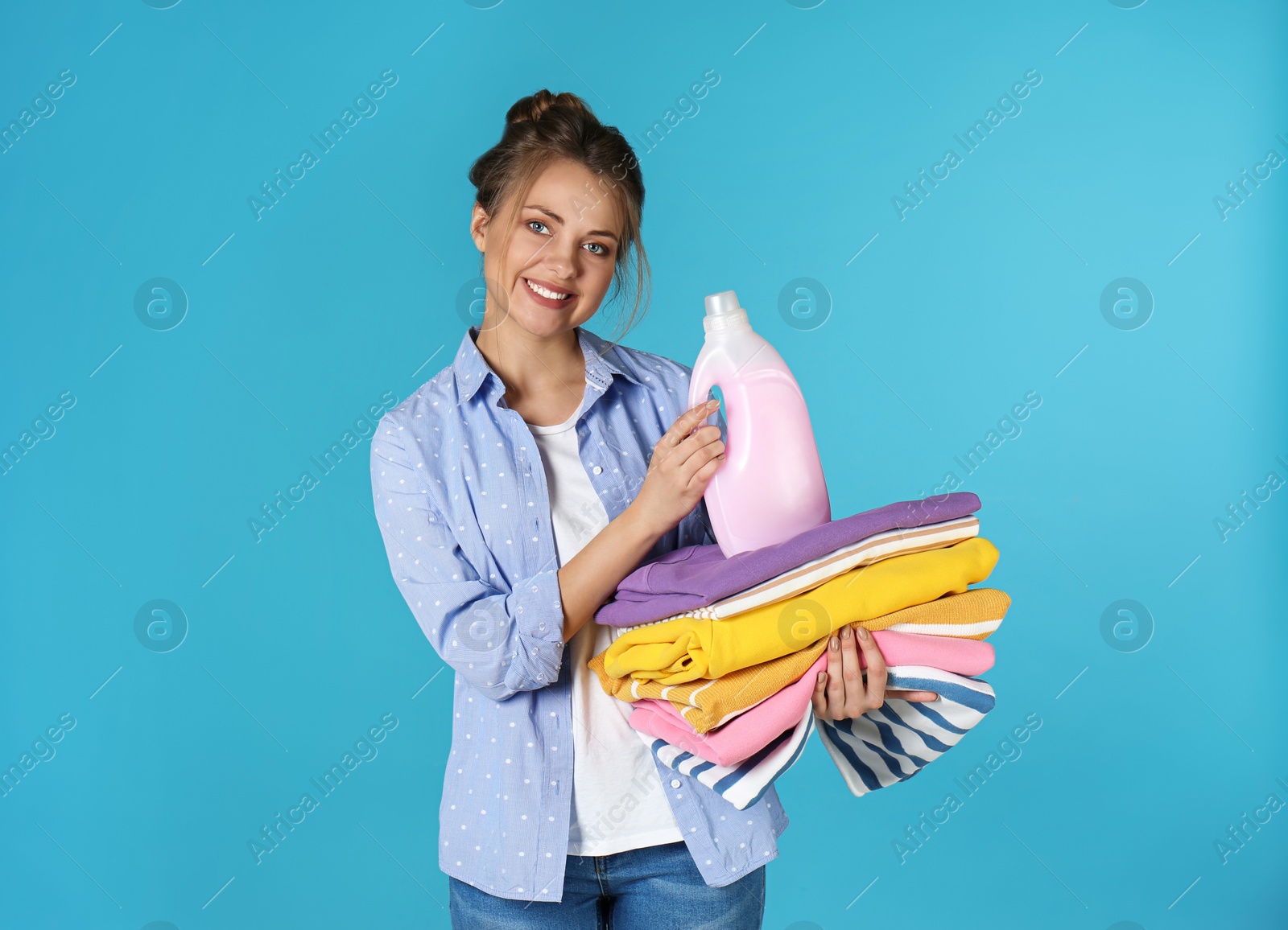 Photo of Happy young woman holding clean clothes and laundry detergent on color background