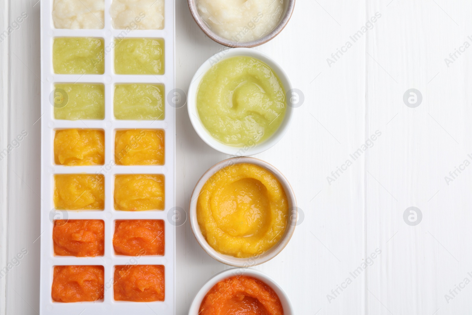 Photo of Different purees in ice cube tray and bowls ready for freezing on white wooden table, flat lay. Space for text
