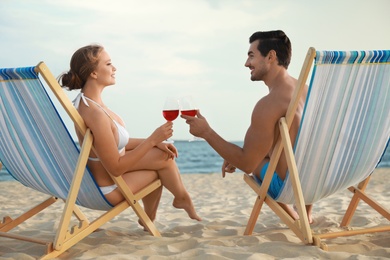 Happy young couple with glasses of wine sitting on deck chairs at sea beach