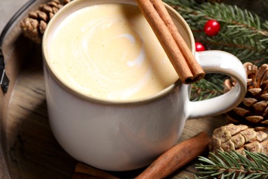 Photo of Tasty eggnog, cinnamon sticks and fir branches on wooden tray, closeup