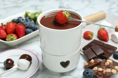Photo of Dipping fresh strawberry in fondue pot with melted chocolate at white marble table