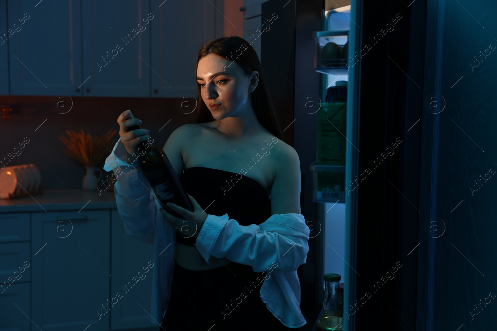 Photo of Beautiful woman with bottle of wine near refrigerator at night