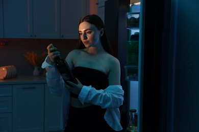 Beautiful woman with bottle of wine near refrigerator at night