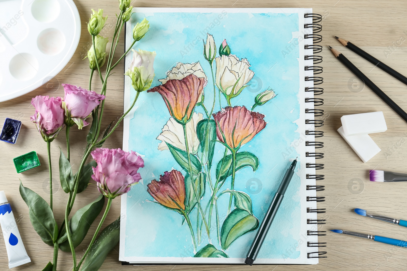 Photo of Painting of eustomas in sketchbook, flowers and art supplies on wooden table, flat lay