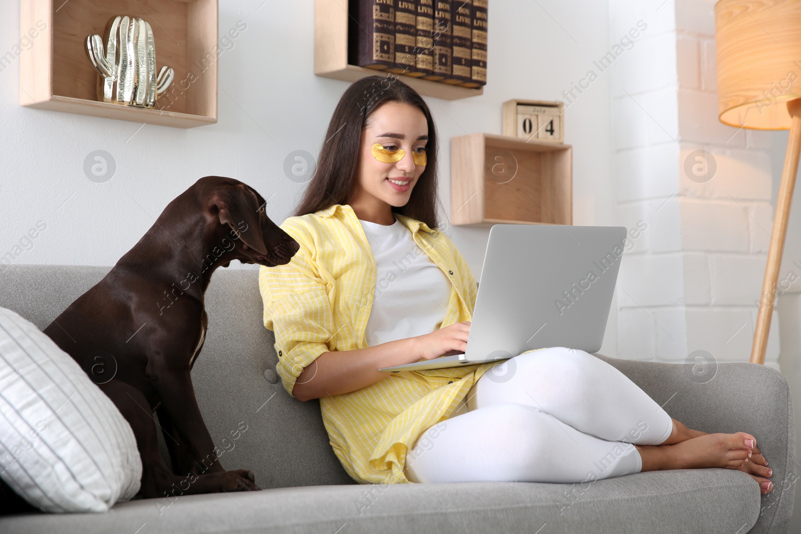 Photo of Young woman with eye patches working on laptop near her dog in living room. Home office concept