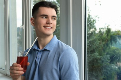 Handsome young man with glass of juice near window at home