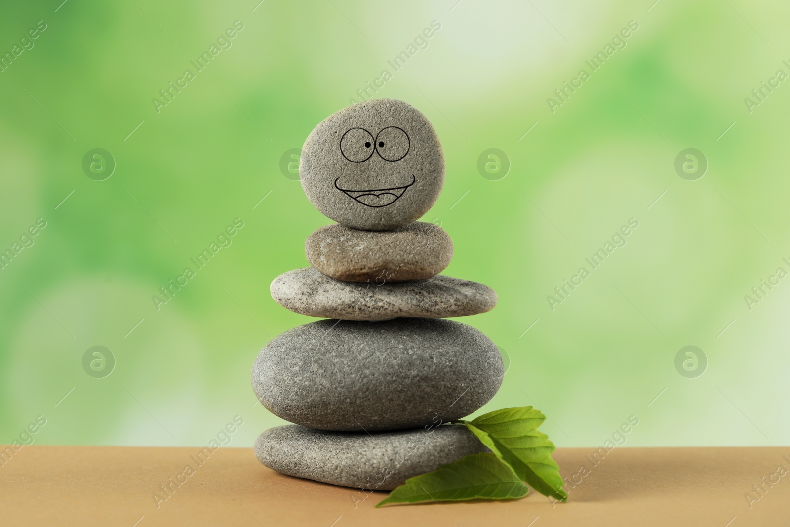 Photo of Stack of stones with drawn happy face and green leaves on table against blurred background. Zen concept