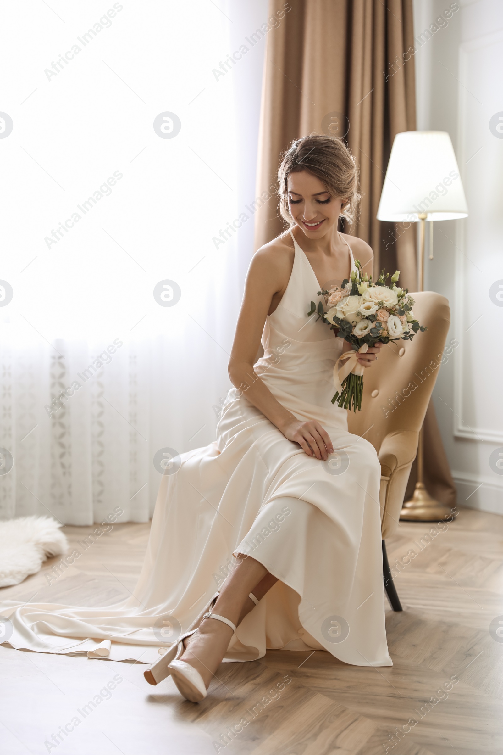 Photo of Beautiful young bride in elegant wedding dress with bouquet sitting in armchair