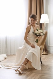 Beautiful young bride in elegant wedding dress with bouquet sitting in armchair