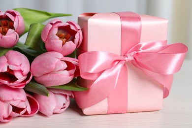 Beautiful gift box with bow and pink tulip flowers on white table, closeup