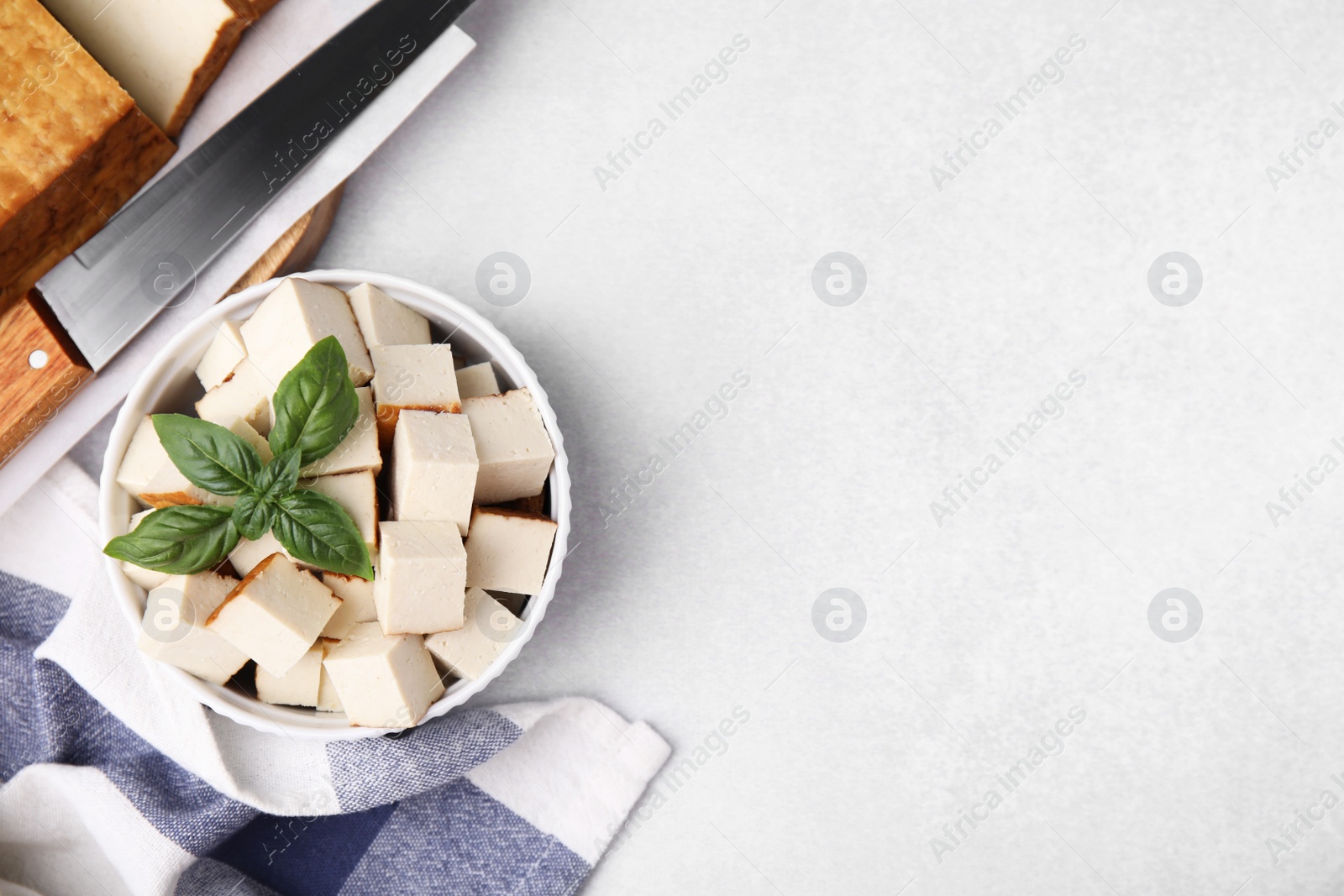 Photo of Bowl of smoked tofu cubes with basil and knife on white table, flat lay. Space for text