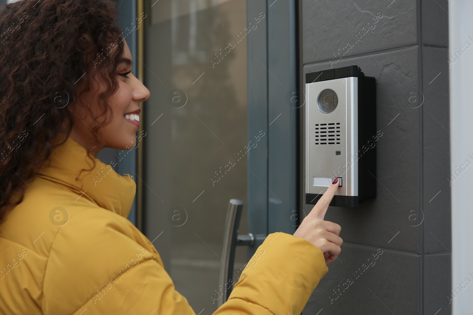 Photo of Young African-American woman ringing intercom with camera near building entrance