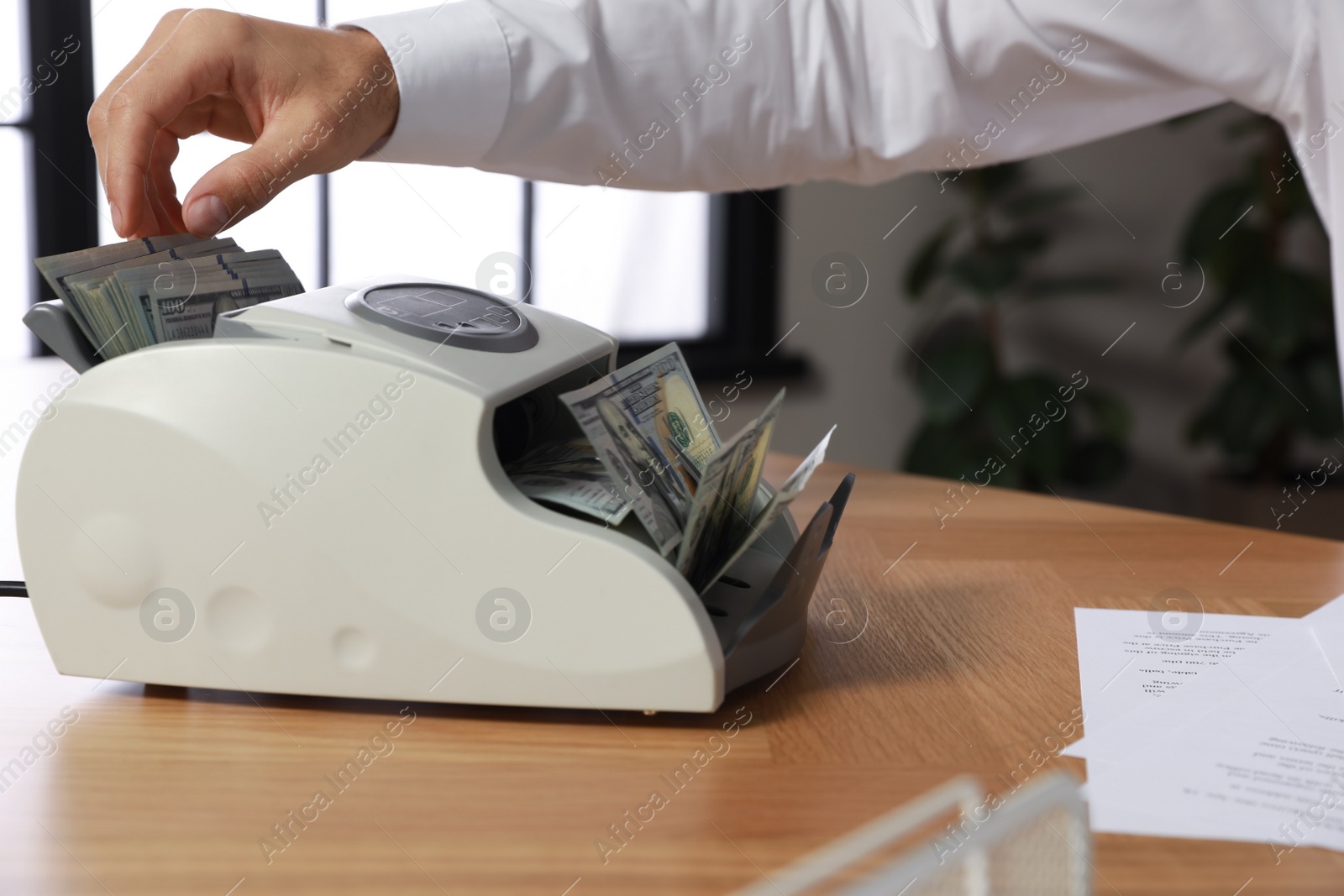 Photo of Man putting money into banknote counter at wooden table indoors, closeup