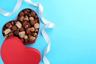 Photo of Different delicious chocolate candies in heart shaped box and ribbon on light blue background, top view. Space for text