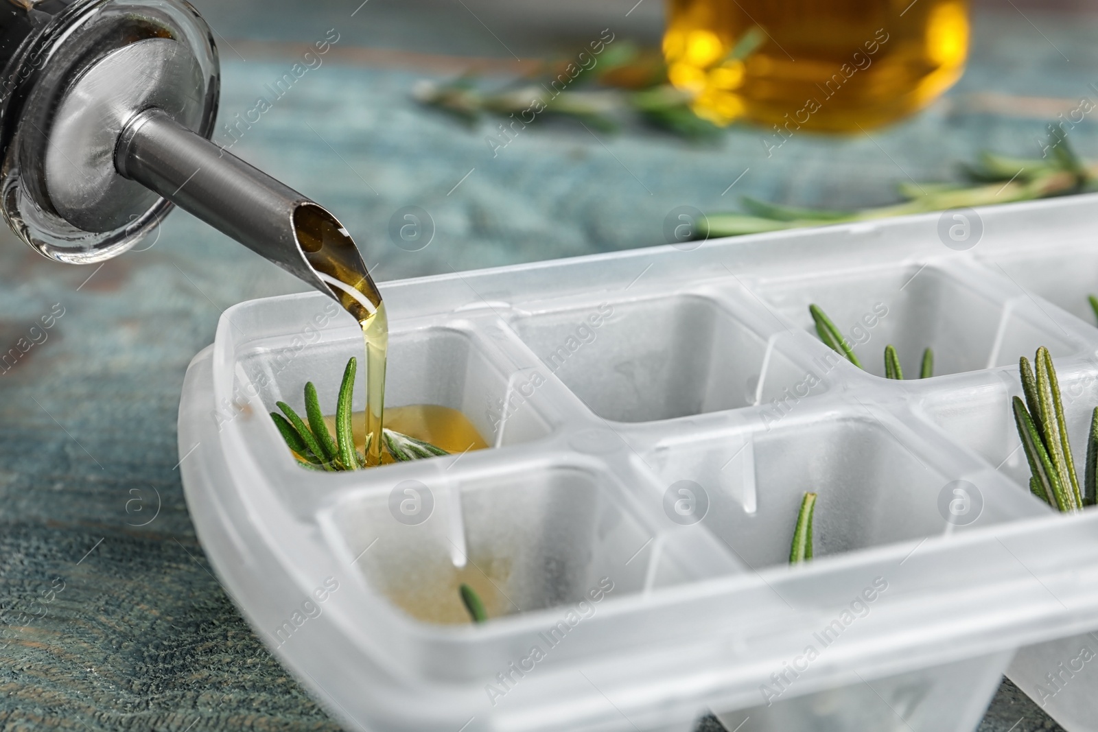 Photo of Pouring olive oil into ice cube tray with rosemary on table, closeup