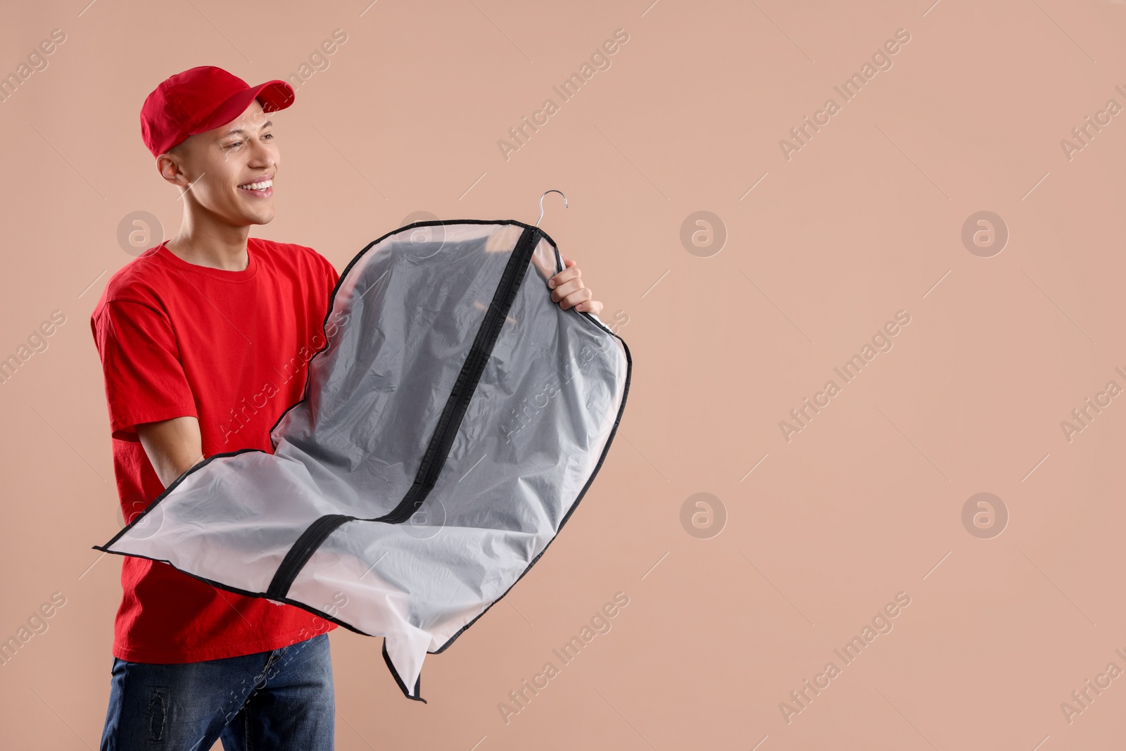 Photo of Dry-cleaning delivery. Happy courier holding garment cover with clothes on beige background, space for text