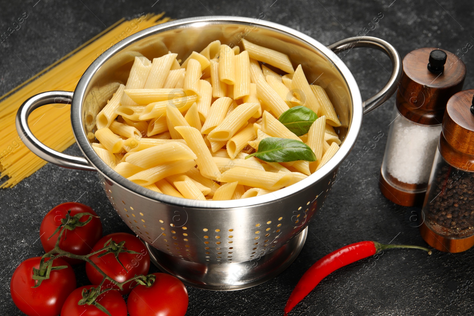 Photo of Cooked pasta in metal colander, products and spices on dark textured table, closeup