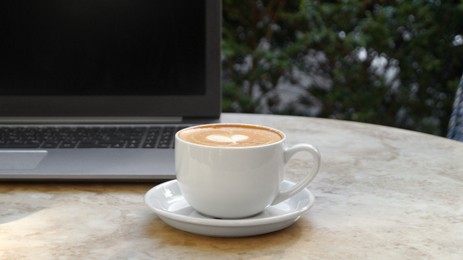 Photo of Cup of delicious coffee and laptop on beige marble table outdoors
